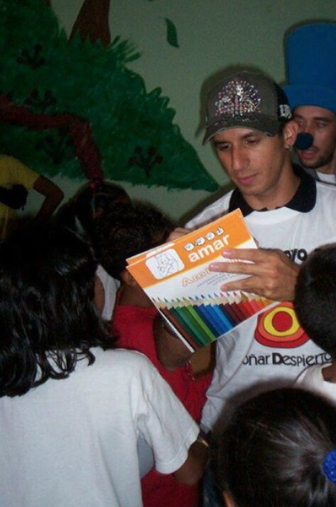 Román EL RO holding a colorful educational gift while surrounded by children.